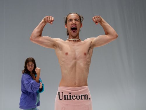 Persona a torso nudo in primo piano posa con le braccia flesse in segno di forza e la bocca spalancata come in un grido. Indossa una collana di perle e un indumento rosa con la scritta “Unicorn.”. Sullo sfondo, leggermente sfocata, un’altra persona con una giacca viola tiene un pugno alzato in una posa combattiva. Lo sfondo è uniforme e chiaro.